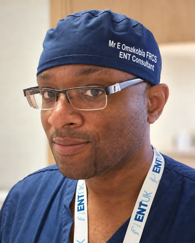 Mr Eugene Omakobia smiling in navy surgical scrubs, cap and glasses