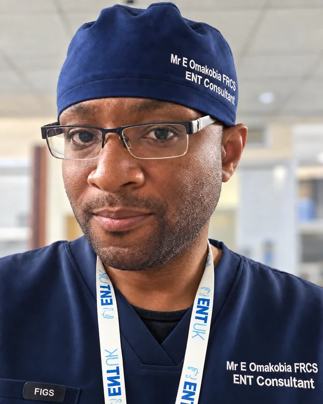 Mr Eugene Omakobia smiling in navy surgical scrubs, cap and glasses
